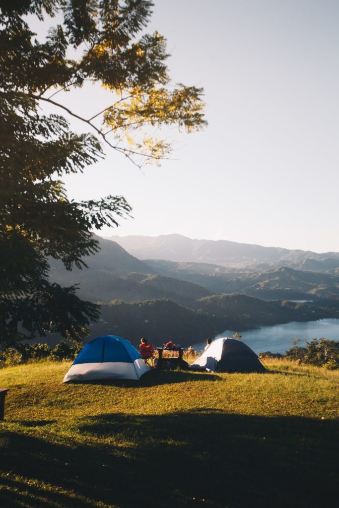 Two tents on a hill overlooking a lake with mountains in the background, with two people sitting outdoors - highlighting the use of bug spray and sunscreen