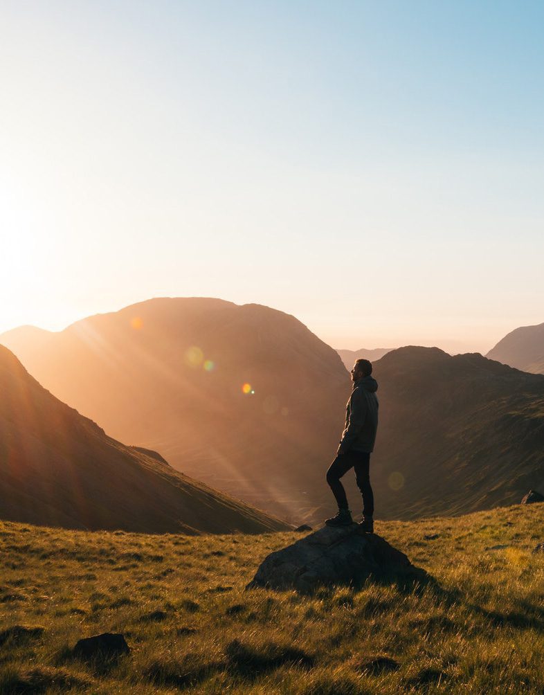 man standing in the sun on a rock on a mountain top at dusk with green grass surrounding